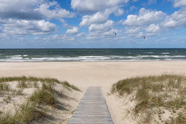 Wooden path through grass-covered Monciškės dunes towards a broad sandy beach, choppy Baltic Sea, and distant kites