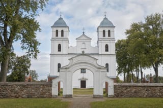 White twin-towered Church of St Michael the Archangel in Mosėdis behind Baroque gates, a fieldstone wall, and red-brick chapels