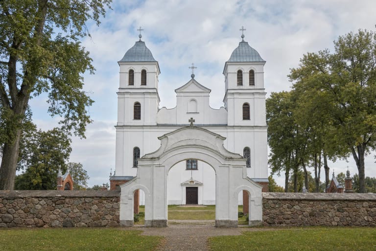 White twin-towered Church of St Michael the Archangel in Mosėdis behind Baroque gates, a fieldstone wall, and red-brick chapels