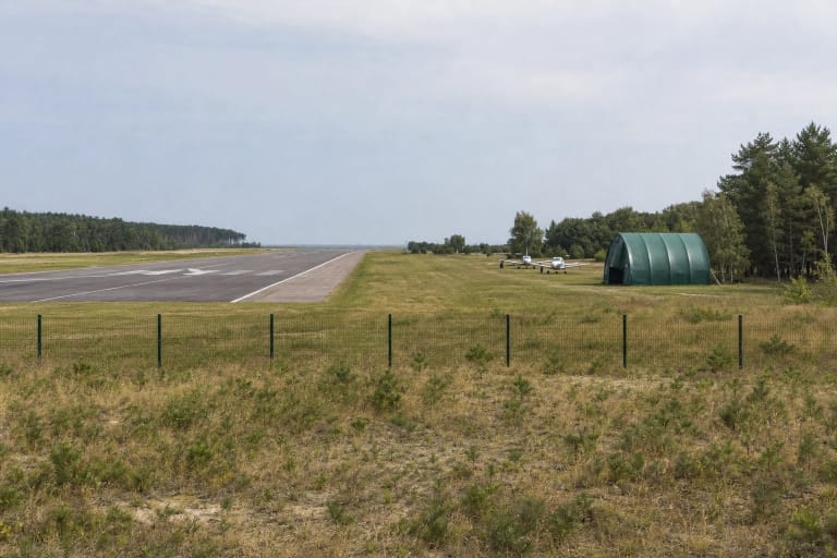 Nidos aerodromo asfaltbetonio takas už žemos metalinės tvoros, apsuptas sausos pievos, pušyno ir Kuršių marių horizonto
