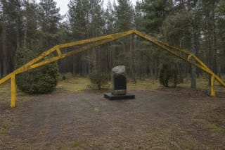 Скромный неровный камень Нидской планёрной школы с тёмной табличкой под жёлтой угловатой аркой ангара на лесной поляне