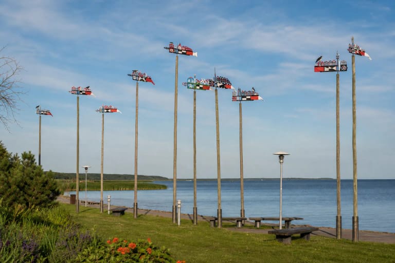 Nidos vėtrungių ekspozicijos aukšti mediniai stiebai su spalvingomis kurėnų vėtrungėmis Kuršių marių krantinėje