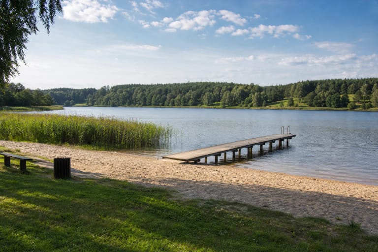 The sandy shore of Orija Lake Beach with a wooden pier, reeds, and forested slopes across the lake