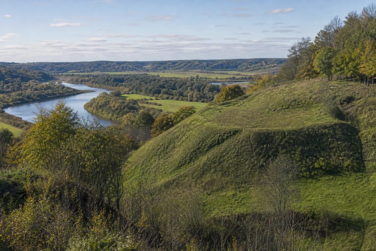 Steep green Padubysys Hillfort with ramparts and deciduous trees above the broad winding Dubysa valley