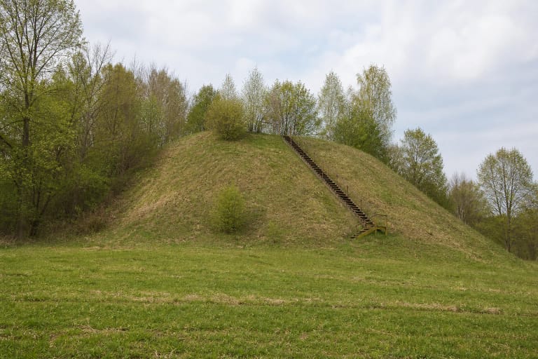 The green cone-shaped Pagaršvys Hillfort seen from the southeast, with timber steps and a belt of deciduous trees