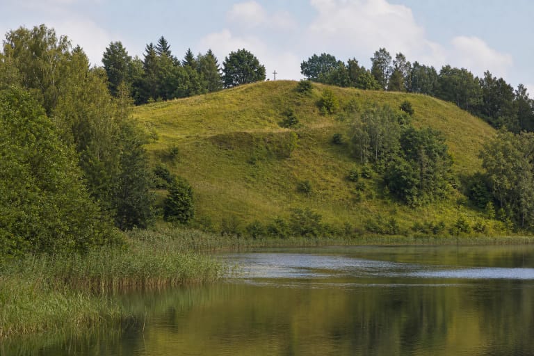 Žolėmis apaugęs Pakalnių piliakalnis prie Vidinksto ežero, apsuptas lapuočių miško