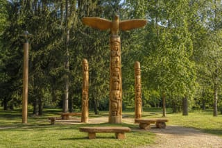 Trys restauruotos Pakruojo miesto parko medžio skulptūros, aukščiausią vainikuoja išskleistų sparnų paukštis, aplink stovi suolai ir brandūs medžiai