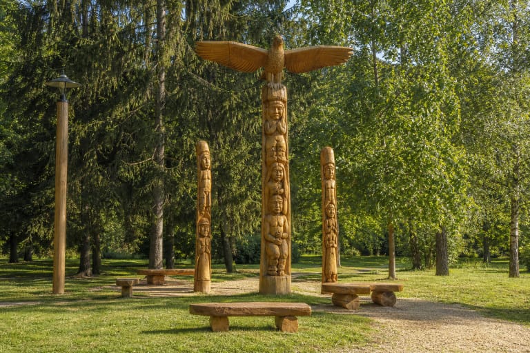 Three restored wood sculptures in Pakruojis City Park, the tallest crowned by a spread-winged bird, with benches and mature trees around them