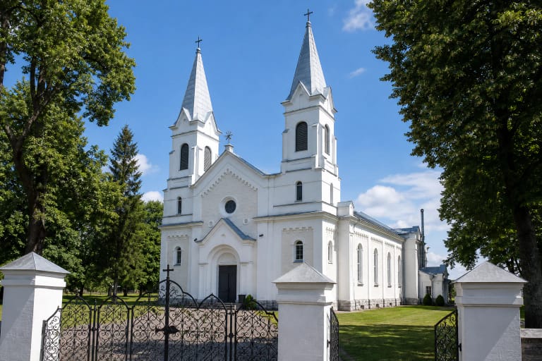 Balta dvibokštė Pakruojo Šv. Jono Krikštytojo bažnyčia su smailiais skardiniais bokštų stogais, apskritu fasado langu ir puošniais šventoriaus vartais