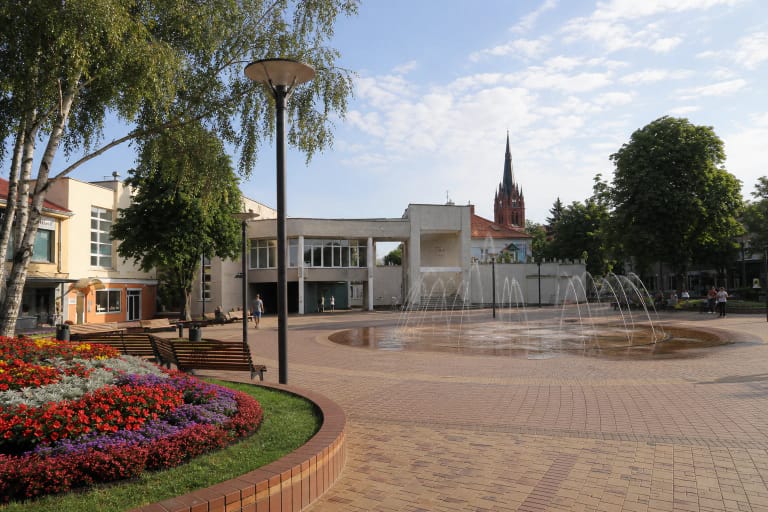 Palangos centrinės aikštės klinkerio grindinys, žemėje įleistas fontanas, spalvingas gėlynas, suolai ir tolumoje matomas bažnyčios bokštas
