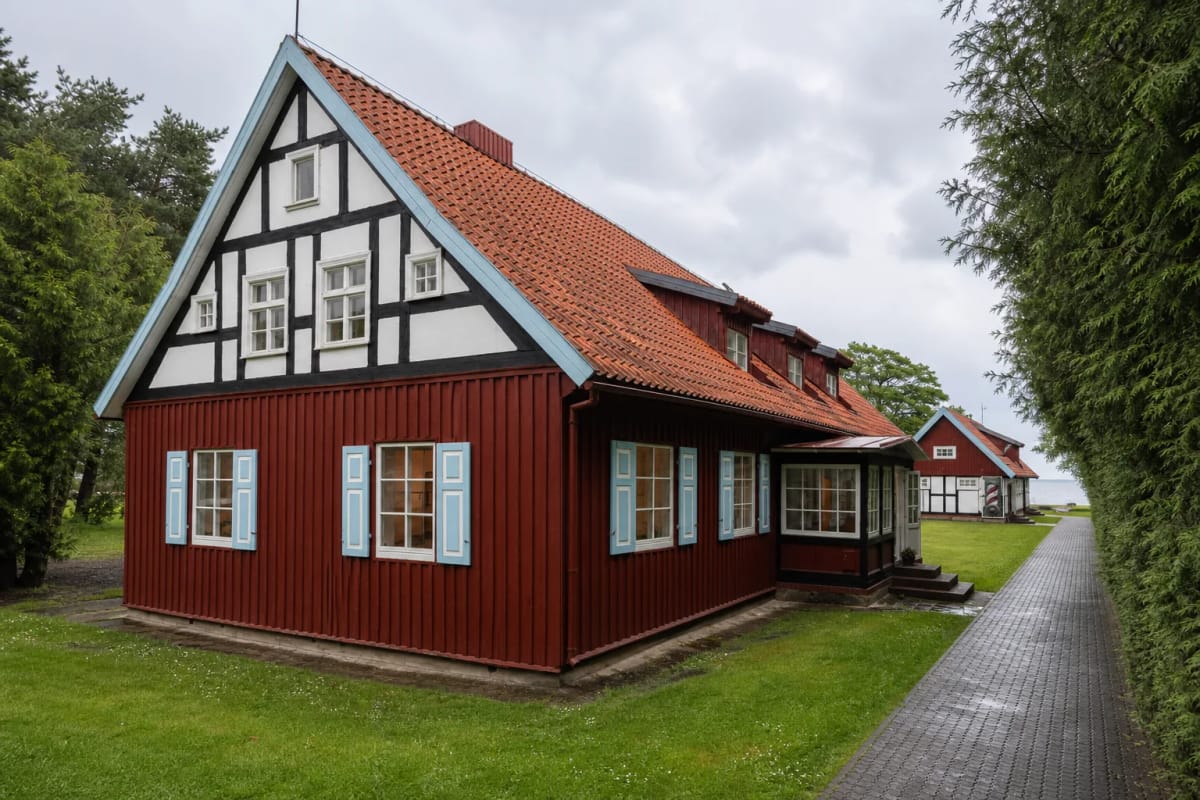 Red timber LNDM Pamario Gallery house with a black-and-white half-timbered gable, pale blue shutters, and tiled roof in Juodkrantė