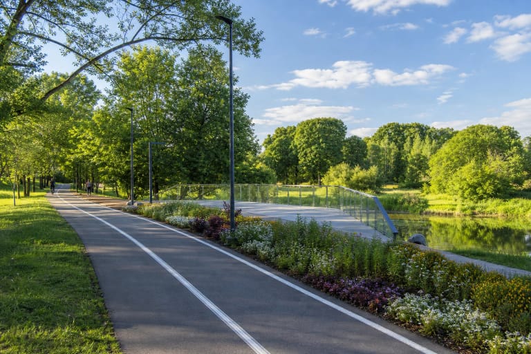 Walking and cycling path in Panevėžys Culture and Recreation Park beside flower beds, mature trees, and a modern bridge over water