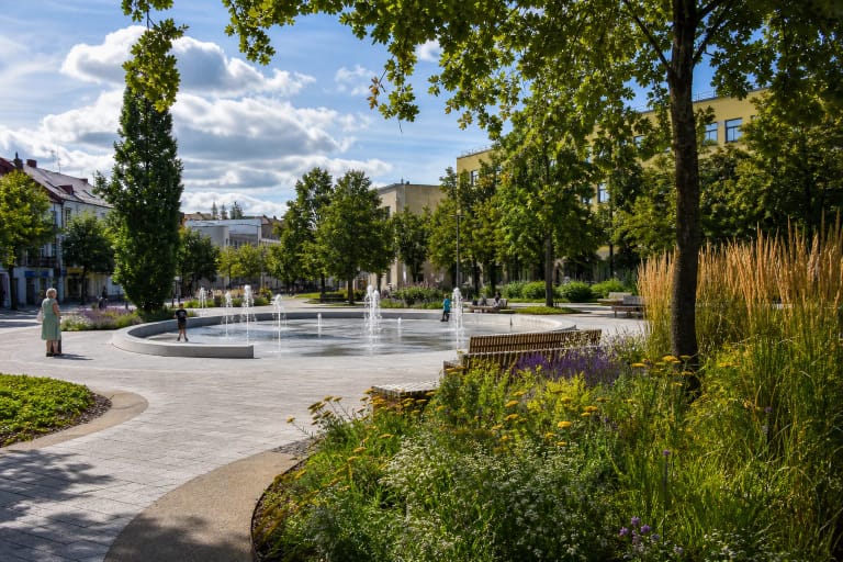 Panevėžio Laisvės aikštės grindinis fontanas tarp gėlynų, brandžių medžių ir miesto centro pastatų