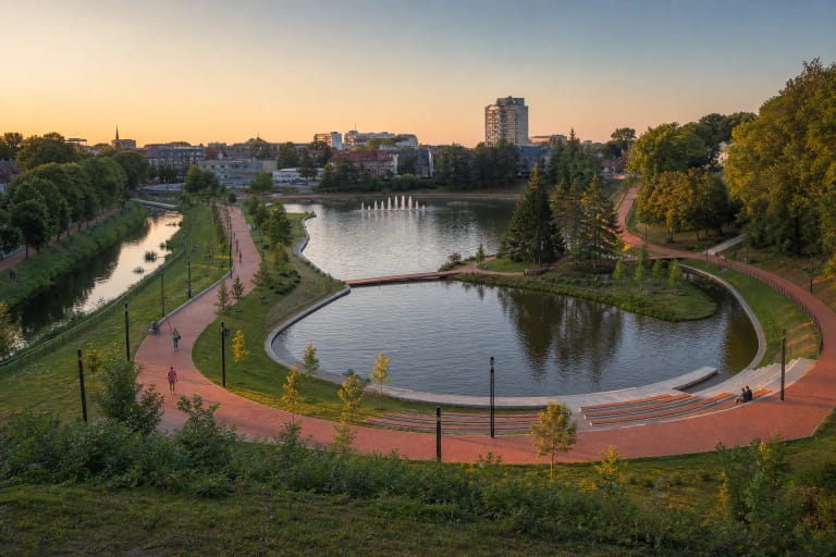 Panevėžio Senvagės tvenkinys, raudonų klinkerio plytų takas, žalia sala ir plaukiojančio fontano srovės vakaro šviesoje