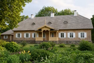 Restored timber house at Pavirvytė Manor with a half-hipped shingle roof, white shutters, and carved porch in a green estate
