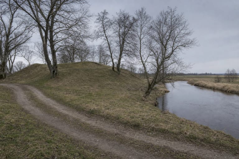 Low grass-covered slope of Piliūnai Hillfort between a bend of the Rausvė and sparse deciduous trees
