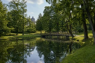 Plungė park pond with a small footbridge, mature trees, and a distant historic roofline