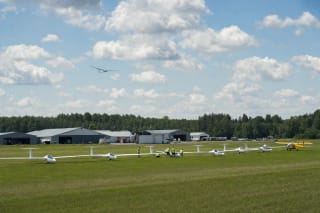 Long-winged white gliders, a yellow tow plane, and ground crew on the grass at Pociūnai Airfield below summer cumulus clouds