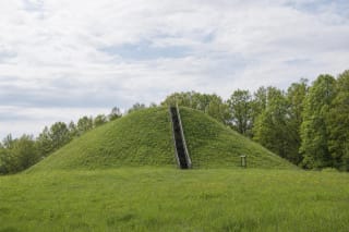 Compact Poteronys Hillfort rising from a spring meadow with long timber steps and trees along the Alovė