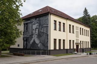 Pale two-storey Prienai Justinas Marcinkevičius Public Library with a brown roof, tall windows, entrance ramp, and monochrome portrait of the poet on its side wall