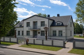 Pale two-storey Prienai Regional Museum with a dark roof, entrance steps, ramp, and carved timber memorial posts in the garden