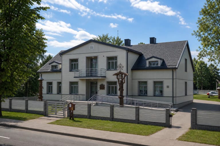 Pale two-storey Prienai Regional Museum with a dark roof, entrance steps, ramp, and carved timber memorial posts in the garden