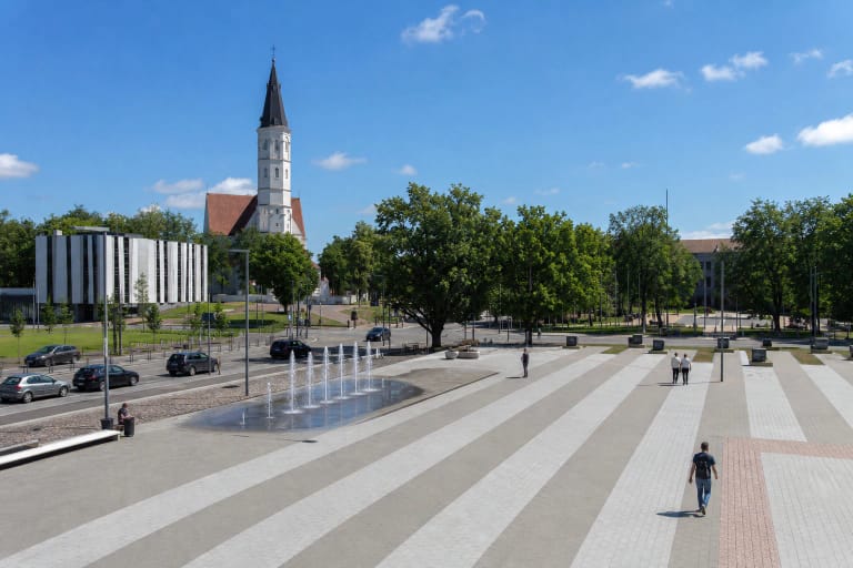 Šviesiomis ir tamsiomis juostomis grįsta Prisikėlimo aikštė su grindiniais fontanais, želdynais ir Šiaulių katedros bokštu fone