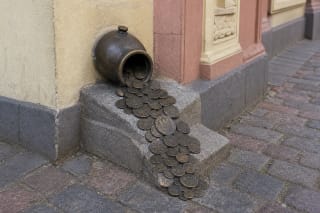 Tipped bronze money pot emerging from the facade at 1 Tiltų Street with coins scattered over a stone ledge