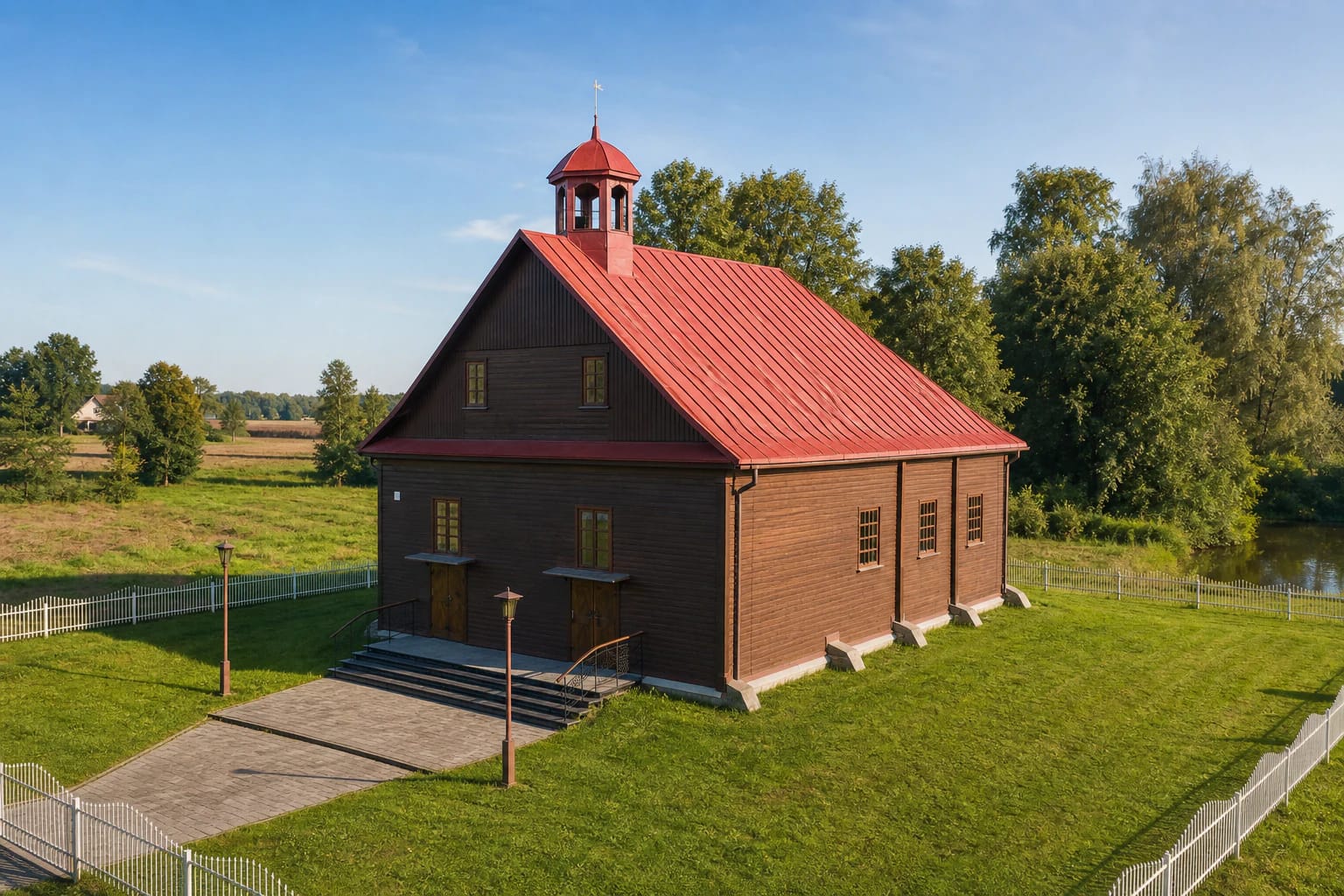 Dark-brown timber walls and red metal roof of Raižiai Mosque, with its crescent turret and white fence beside a green pond