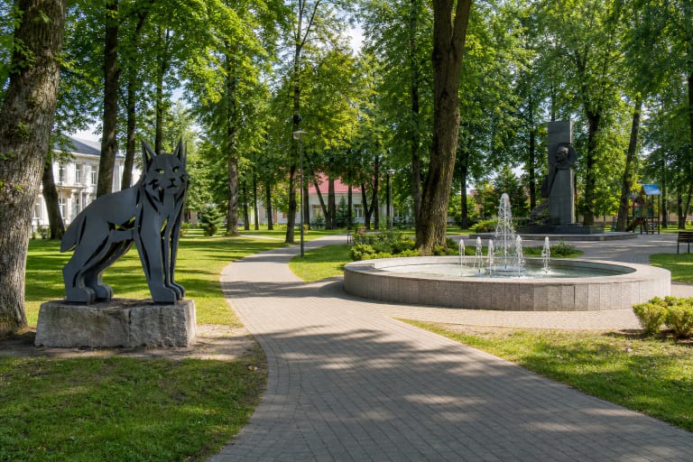 Green path in Raseiniai Maironis Park leading past a fountain and contemporary lynx sculpture among mature trees
