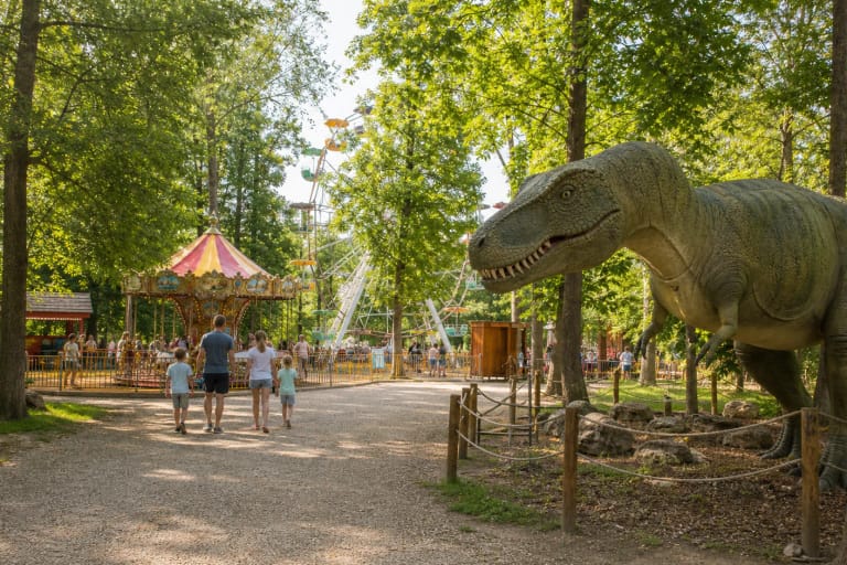 Raubonių parko miško take stovintis didelis dinozauro modelis, o fone matyti karuselė, apžvalgos ratas ir šeimos