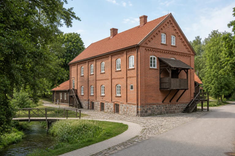 Restored red-brick Raubonys watermill-cardery-spinnery with a tiled roof, timber hoist balcony, and footbridge over a narrow channel