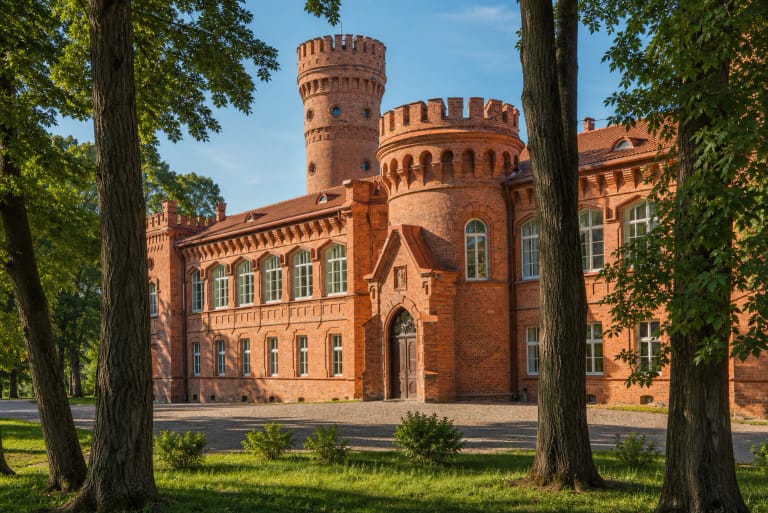Raudonės pilies raudonų plytų fasadas ir bokštai matomi pro parko medžius