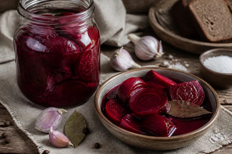 Fermented beets in a glass jar with dark red rasalas, a slice of rye bread, garlic, and a wooden spoon