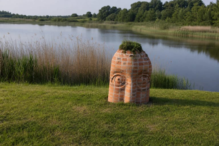 Rokai Recreation Area shore with a two-faced brick marker, grass, reeds, and a Paraželiai pond