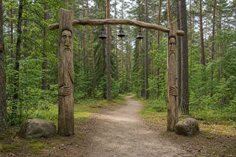 Carved gateway of Rozalimas Forest Park with three bells above a sandy path through green pine woodland