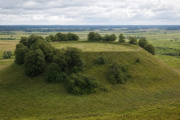 The grassy ring bank and lime-fringed enclosure of Rudamina Hillfort above wet Dzūkija meadows in late summer