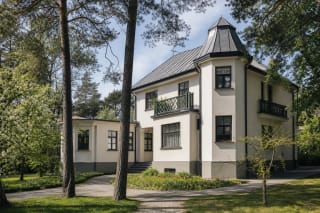 Белая вилла Саломеи Нерис и Бернардаса Бучаса с тёмной крышей и зелёным балконом среди сосен Палемонаса