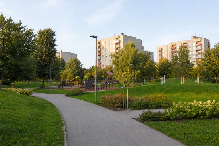 A curving asphalt path, lawn, children's playground, and Šilainiai apartment blocks among the trees in Santarvė Park