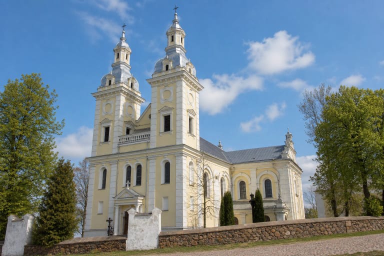 Šviesi Šeduvos Šv. Kryžiaus Atradimo bažnyčia su dviem barokiniais bokštais už akmenų mūro šventoriaus tvoros