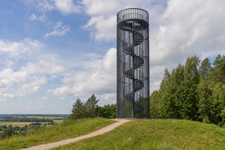 Cilindrinis Šiaulės kalno apžvalgos bokštas su spiraliniais laiptais ant žalios kalvos Gomertos kraštovaizdyje