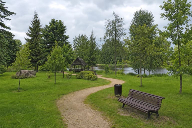 A winding pale gravel path through Šilalė Community Park, bordered by lawn, young trees, timber benches, and the town pond