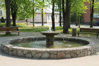 Apskritas akmeninis Šilutės fontanas su centrine taure ir vandens čiurkšlėmis medžių apsuptame skvere