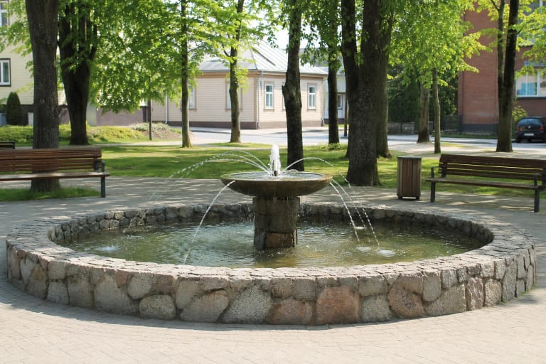 Apskritas akmeninis Šilutės fontanas su centrine taure ir vandens čiurkšlėmis medžių apsuptame skvere
