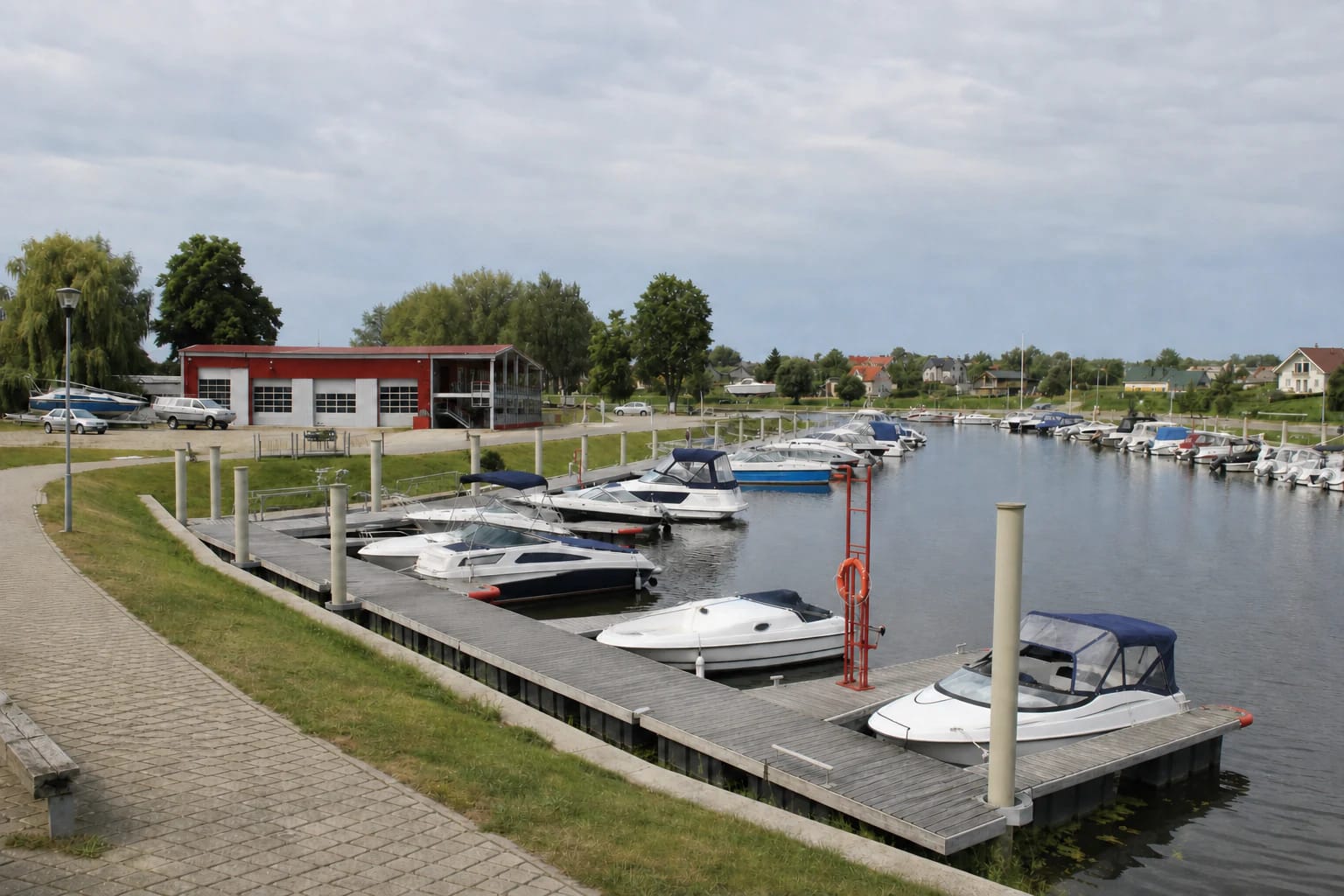 Šilutė Small-Craft Harbour basin with grey floating finger piers, modest motorboats, and the red harbour building in a Šyša oxbow