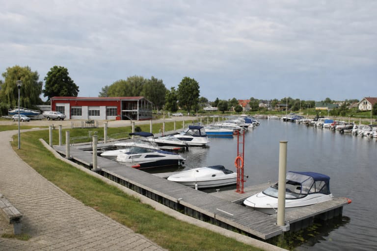 Šilutės mažųjų laivų uosto baseinas su pilkais pontoniniais lieptais, nedideliais kateriais ir raudonu uosto pastatu Šyšos senvagėje