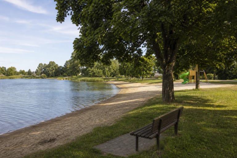 The sandy Širvintos Beach shore by the pond with tree shade, a bench, and a green slide