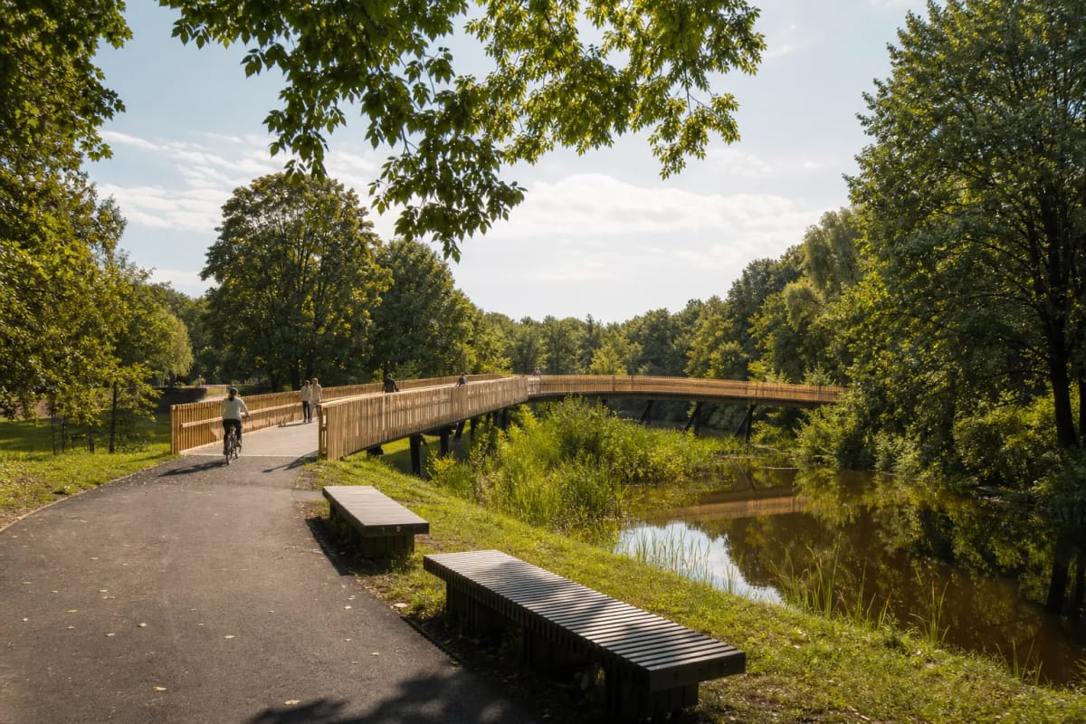 Pale-timber pedestrian bridge over a Skaistakalnis Park pond among mature trees and asphalt paths