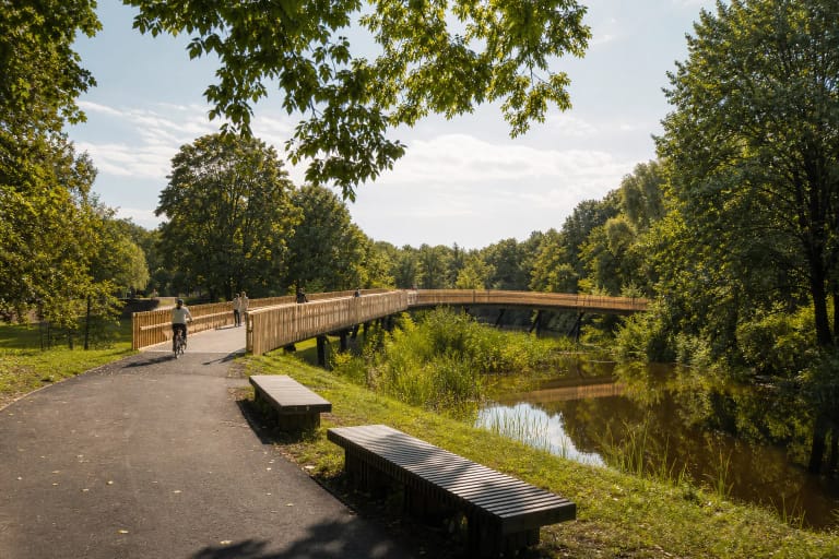 Pale-timber pedestrian bridge over a Skaistakalnis Park pond among mature trees and asphalt paths