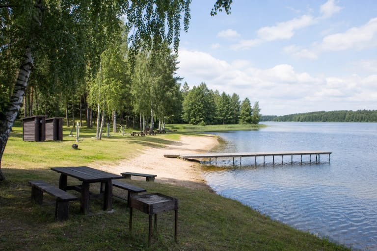 Skaistė Lake Beach with a sandy shore, pier, picnic tables, and trees